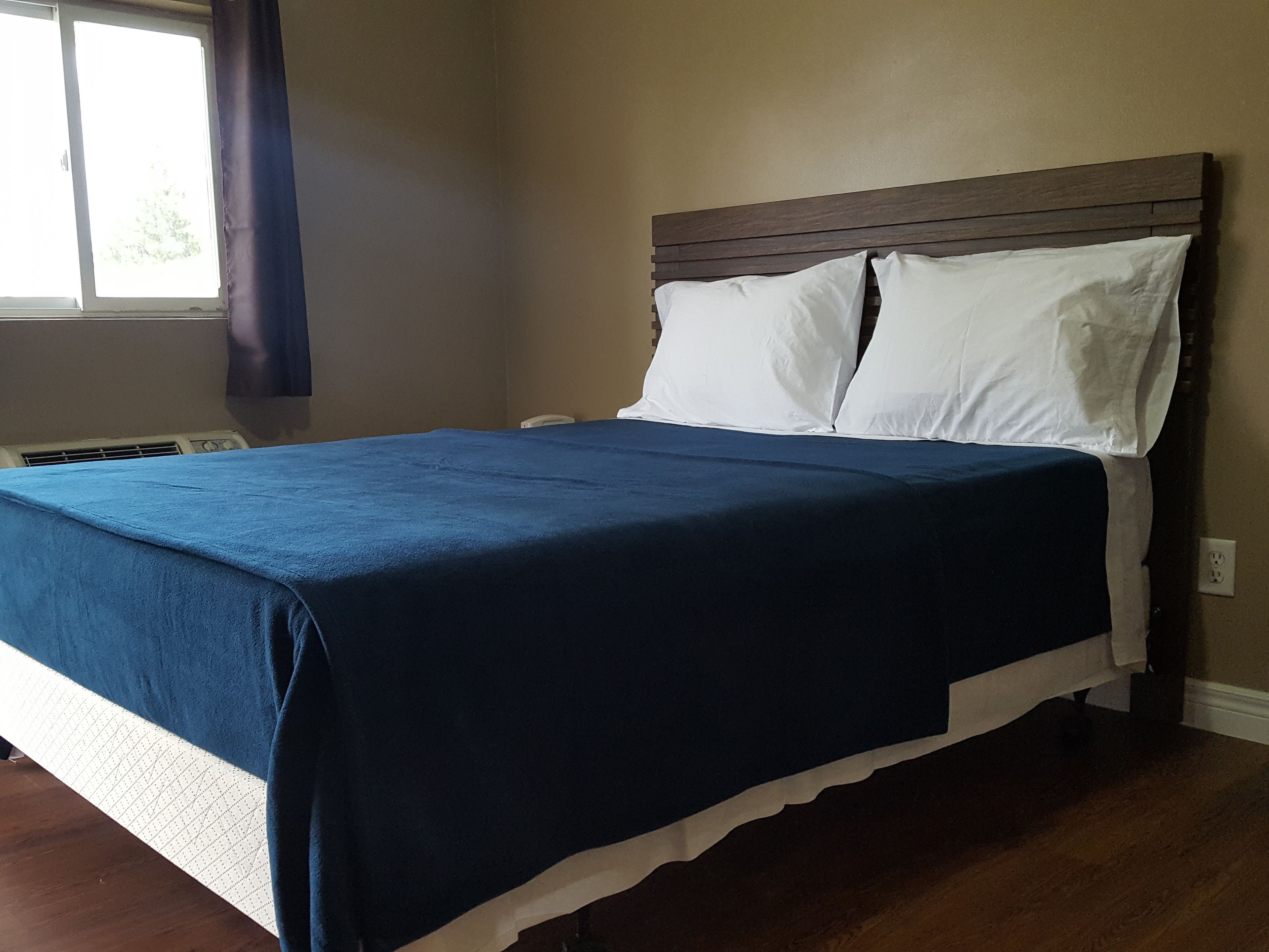 Un lit bien fait, avec une couvre-lit bleue et des oreillers blancs, une tête de lit en bois et une fenêtre avec des rideaux.