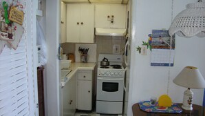 A compact kitchen with white cabinets, a stove, and a microwave. There is a calendar on the wall and a small table with a lamp. - Ocean Suite Beach Front at Mobay Club (Montego Bay)