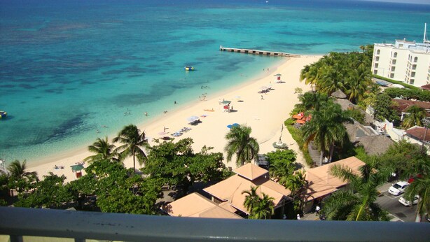 A beach with clear turquoise water, a pier extending into the sea, and a hotel with a white facade and multiple windows. - Ocean Suite Beach Front at Mobay Club (Montego Bay)