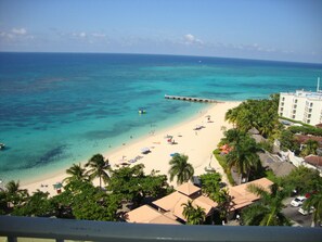 A beach with clear turquoise water, a pier extending into the sea, and a hotel with a white facade and multiple windows. - Ocean Suite Beach Front at Mobay Club (Montego Bay)