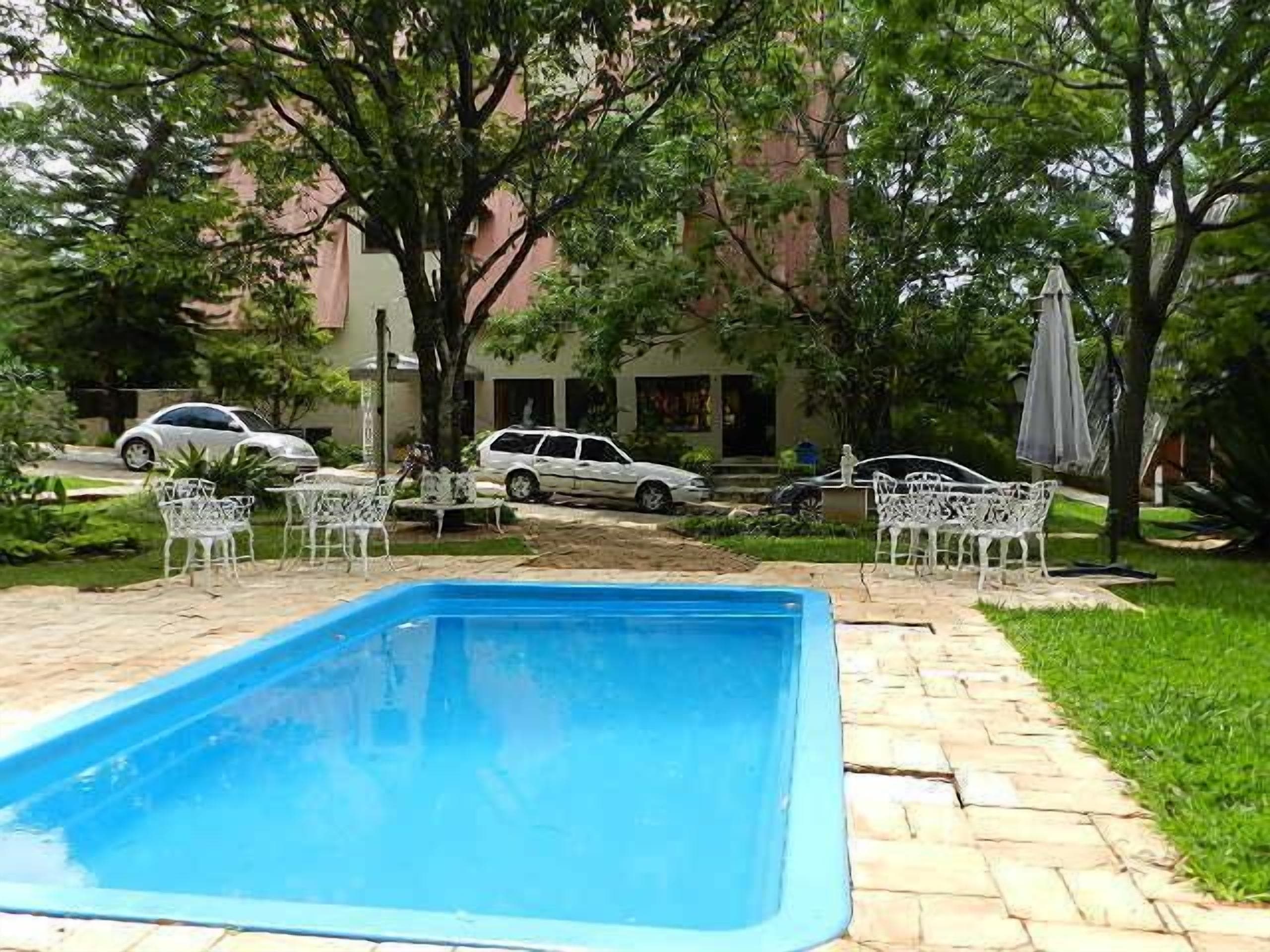 Piscina com deck pavimentado, rodeada por cadeiras e mesas de metal branco, com árvores e um edifício ao fundo.