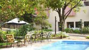 An outdoor pool area with lounge chairs and umbrellas, surrounded by greenery and a building in the background. - Chalé Hotel (Bauru)