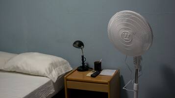 En seng med hvide sengetøj, et natbord med lampe, fjernbetjening, en kop og en gulvventilator.