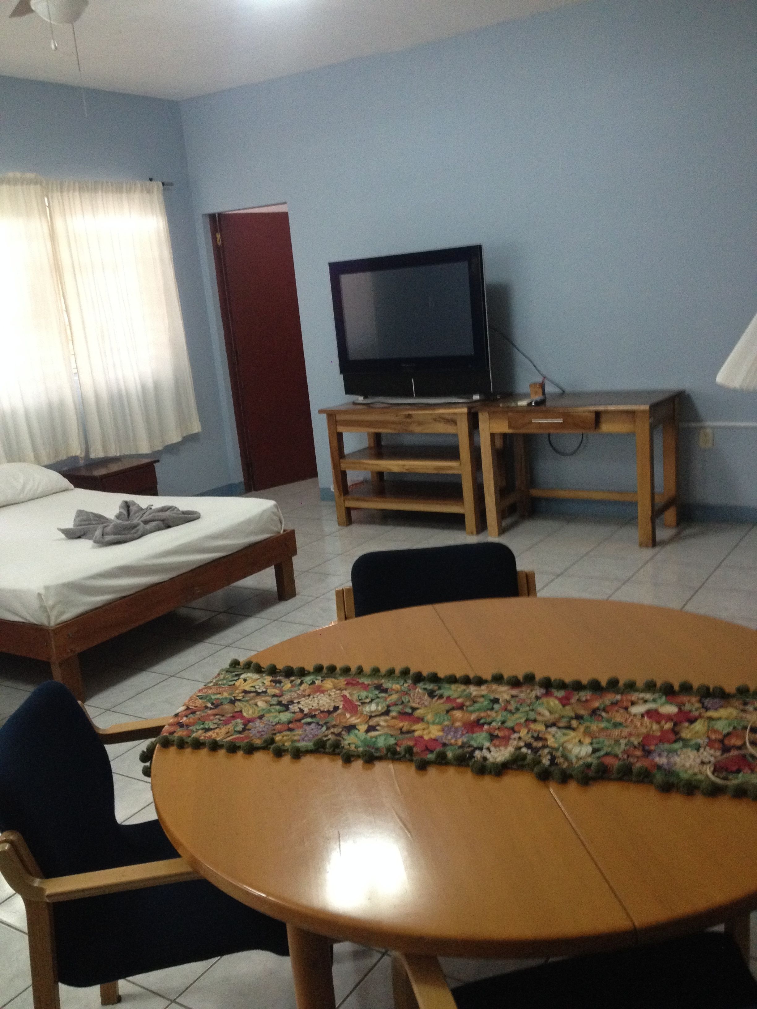 A room with a bed, a TV on a wooden stand, a round table with a colorful table runner, and a chair.