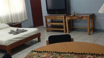 A room with a bed, a TV on a wooden stand, a round table with a colorful table runner, and a chair.