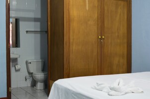 Una habitación de hotel con un armario de madera, una cama blanca con toalla blanca, un inodoro blanco y un lavamanos pequeño.