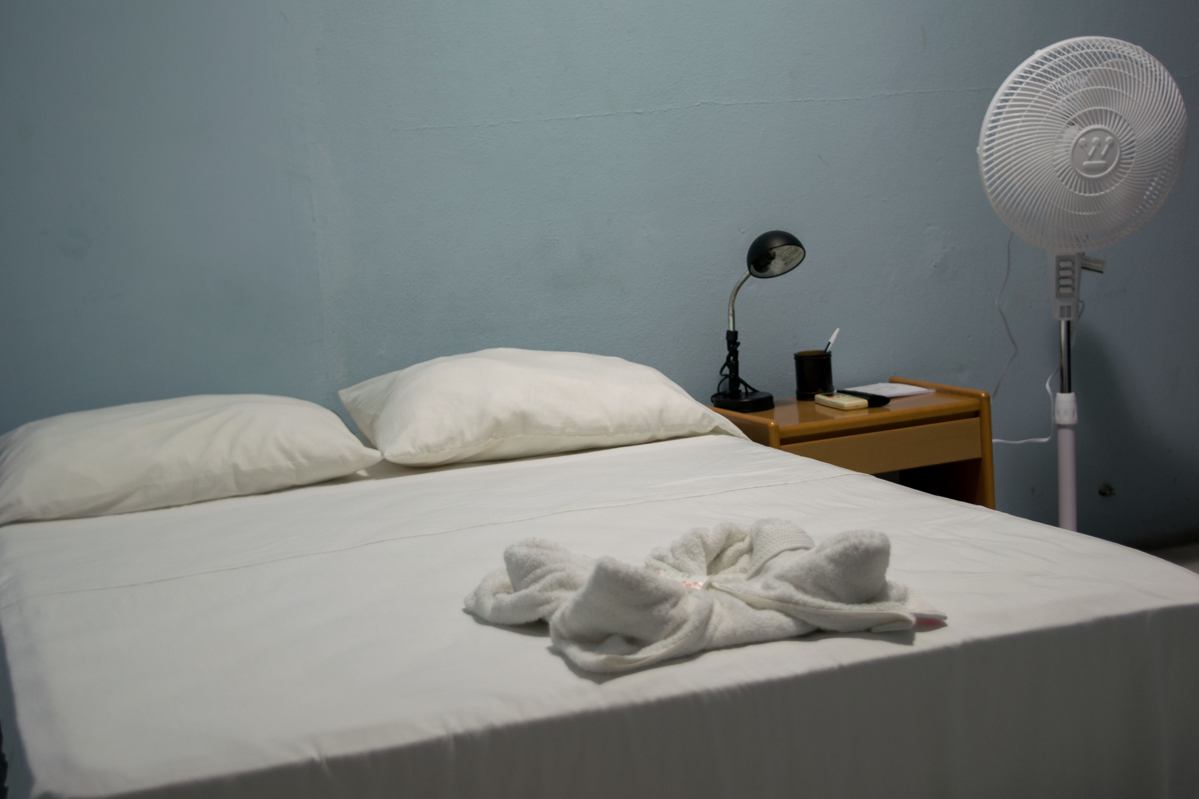 Una habitació d'hotel amb un llit, almoines, una taula de nit amb una làmpada i un ventilador de peu.