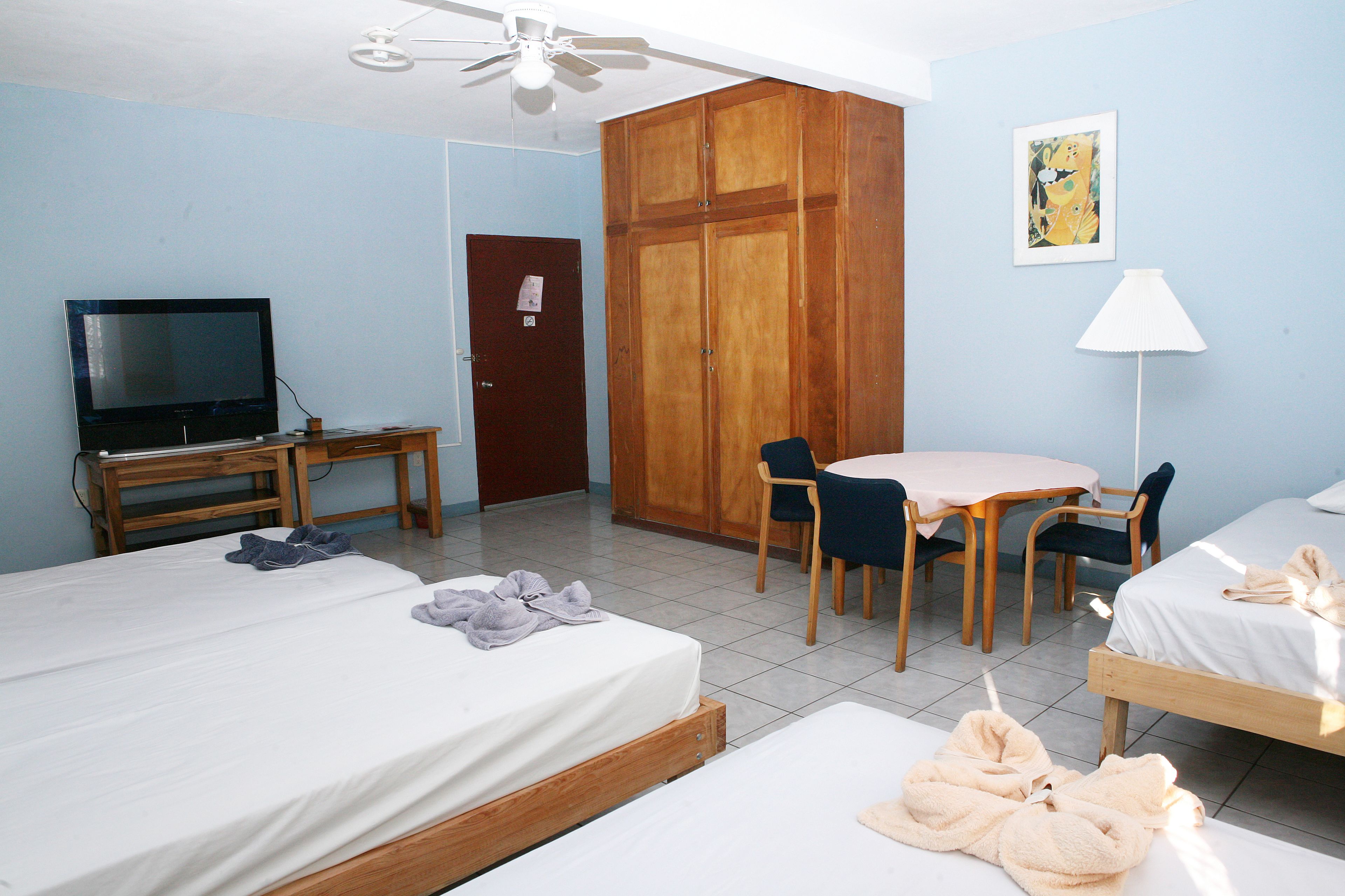 A hotel room with two beds, a TV, a wardrobe, a dining table with chairs, and a ceiling fan.