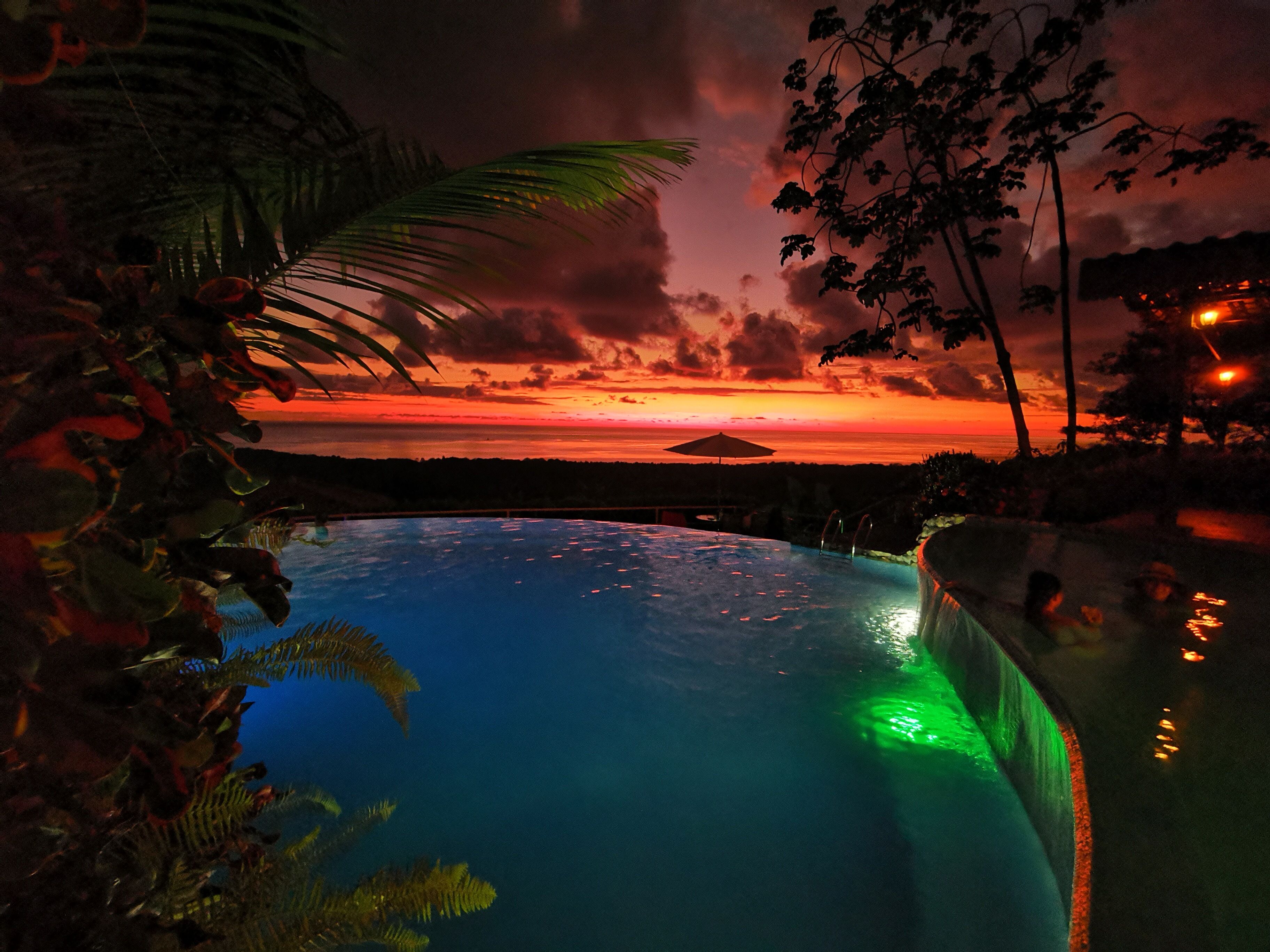 Ein Pool mit Blick auf einen Sonnenuntergang über dem Meer, umgeben von Palmen und einem Strand.
