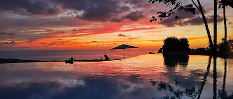 An infinity pool reflecting a sunset with a beach view, palm trees, and a gazebo.
