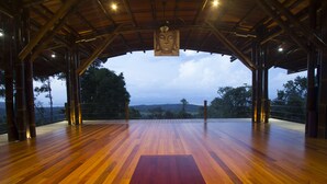 Un espace ouvert avec un plancher en bois, agrémenté d’un tapis rouge au centre, d’une statue de Bouddha suspendue et offrant une vue sur les arbres et les collines à travers une zone abritée.