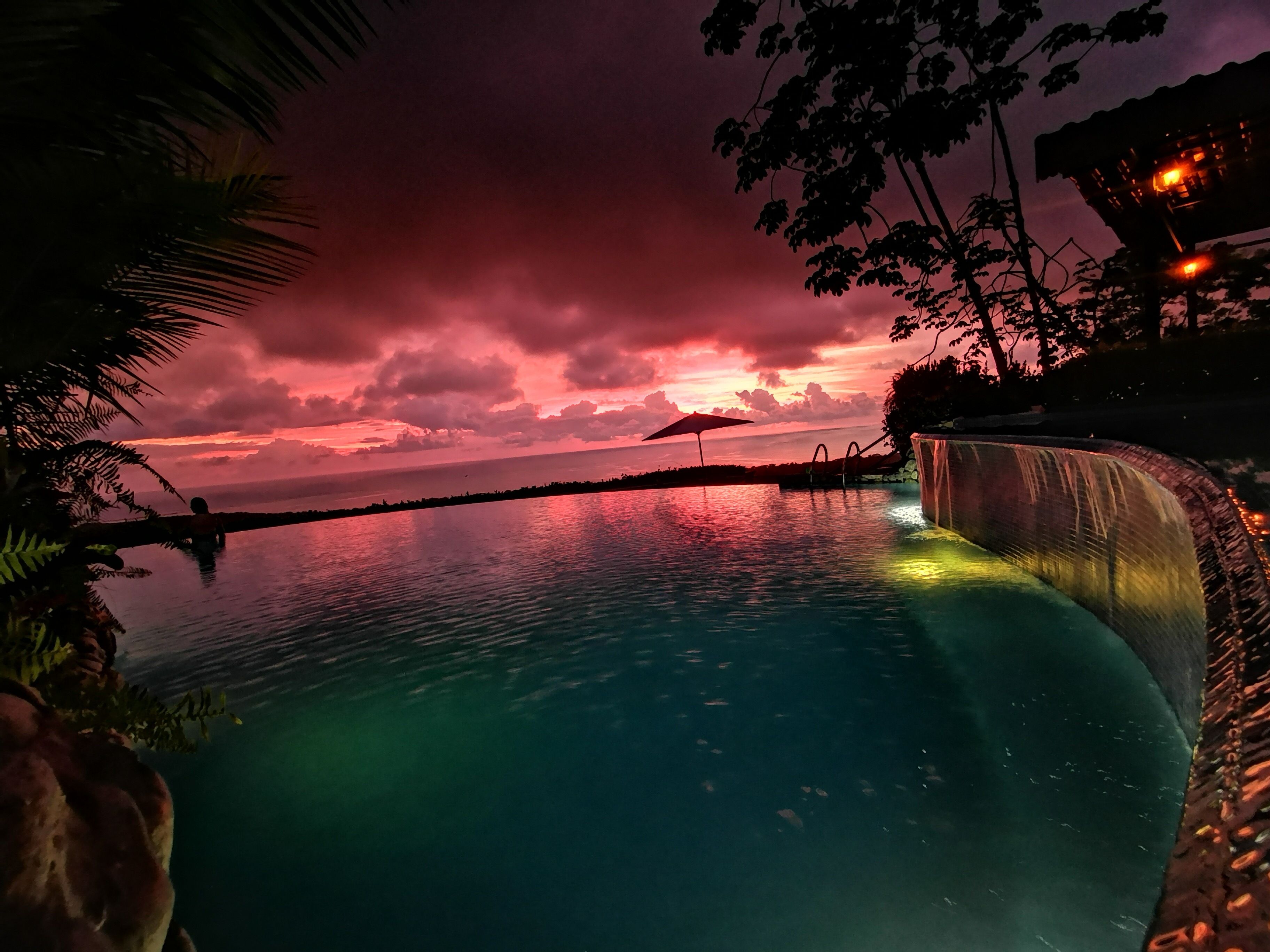 A serene pool area with a waterfall feature, surrounded by lush greenery and a sunset sky.