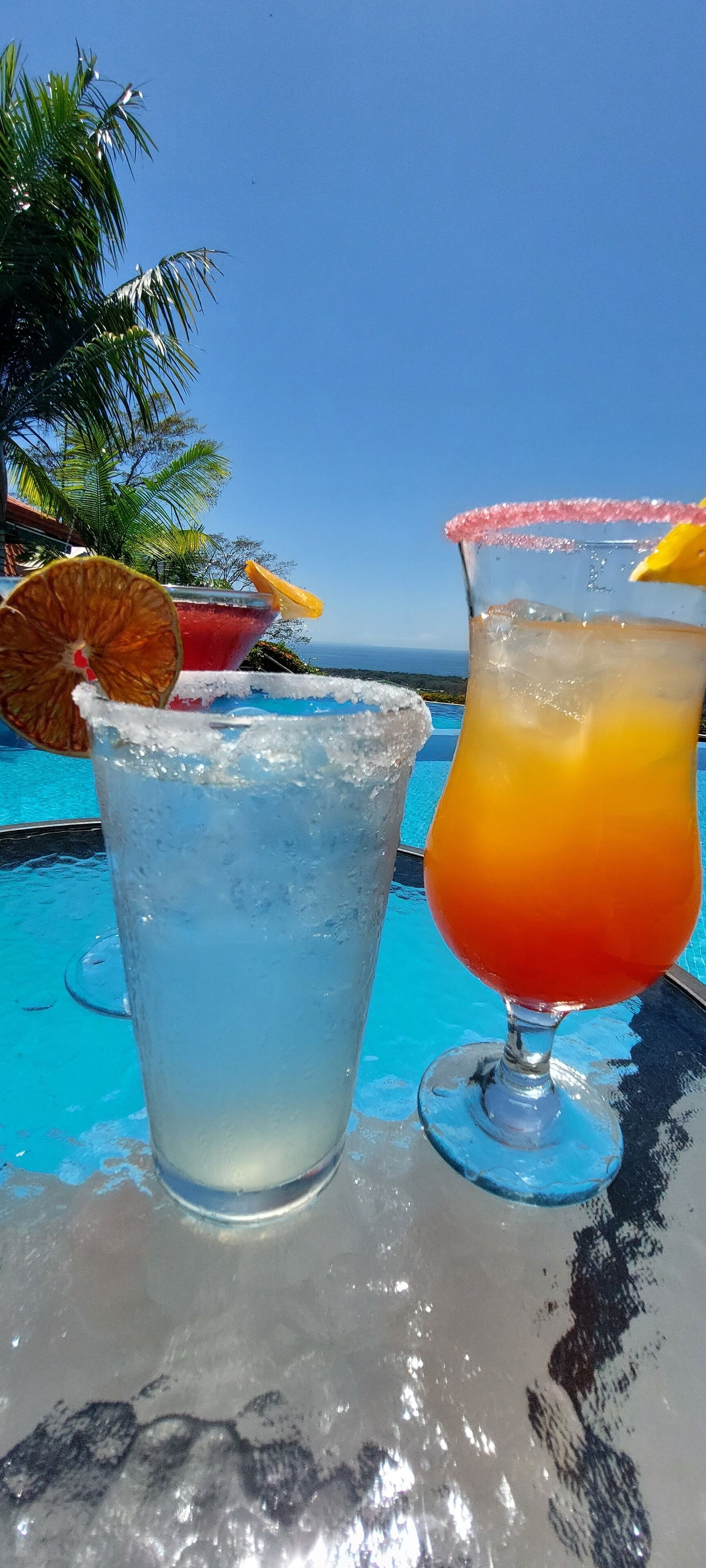 Deux boissons sur une table près d’une piscine, avec un ciel dégagé et des palmiers en arrière-plan.
