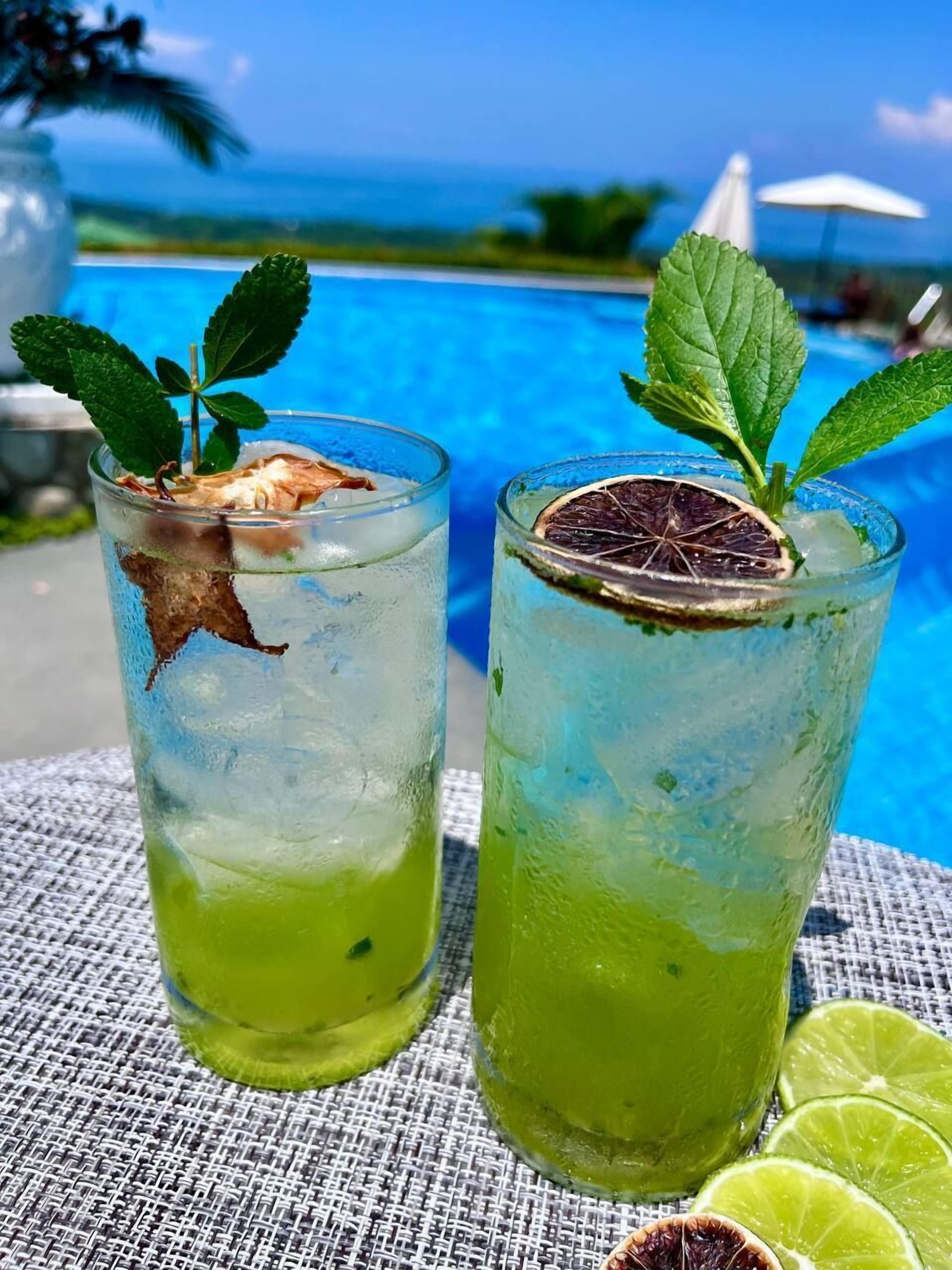 Dos vasos de cóctel verde con hojas de menta y una guarnición de fruta seca, sobre una mesa con vista a la piscina.