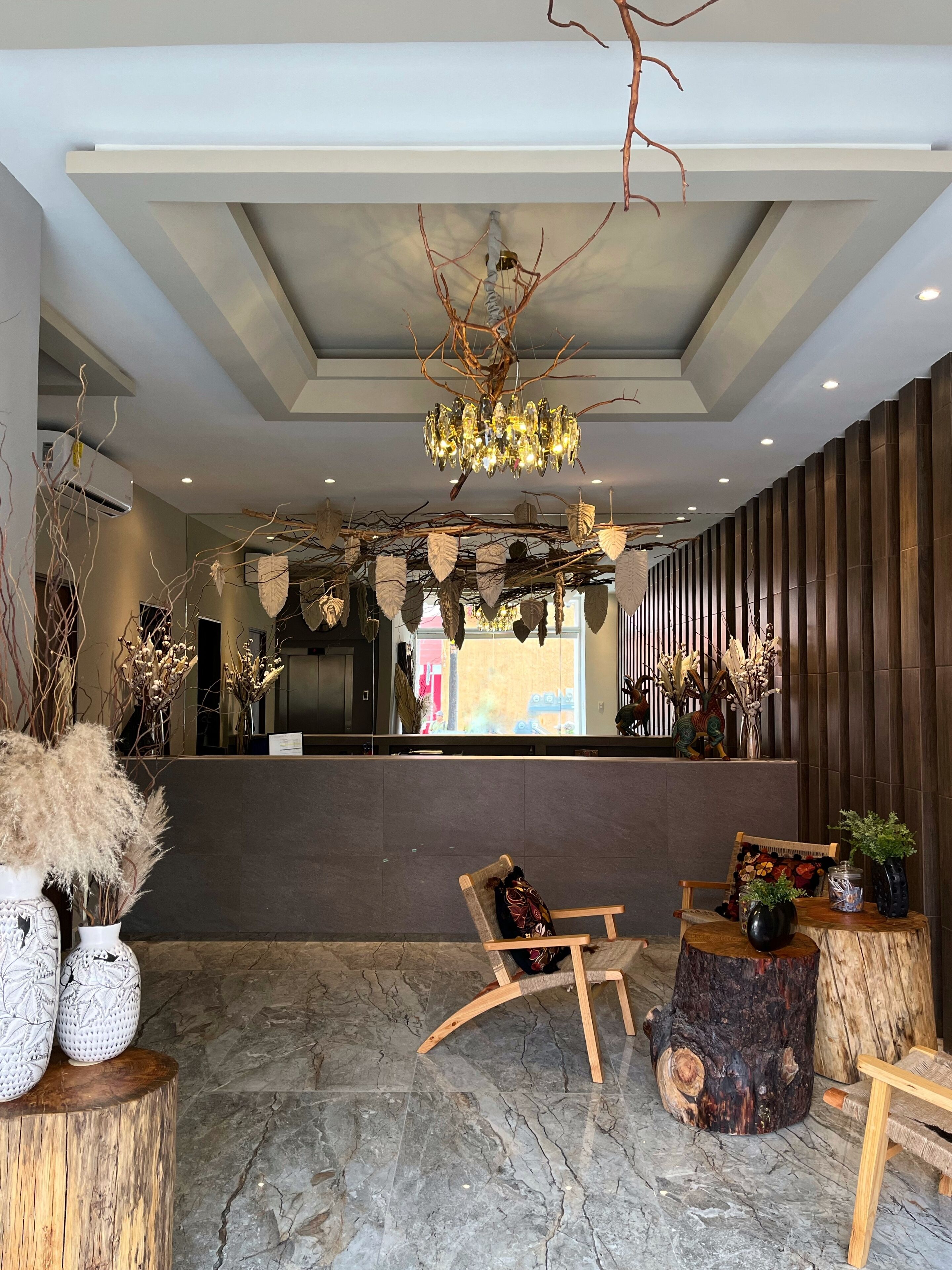 A lobby with a modern chandelier, wooden reception desk, seating area, and decorative plants.