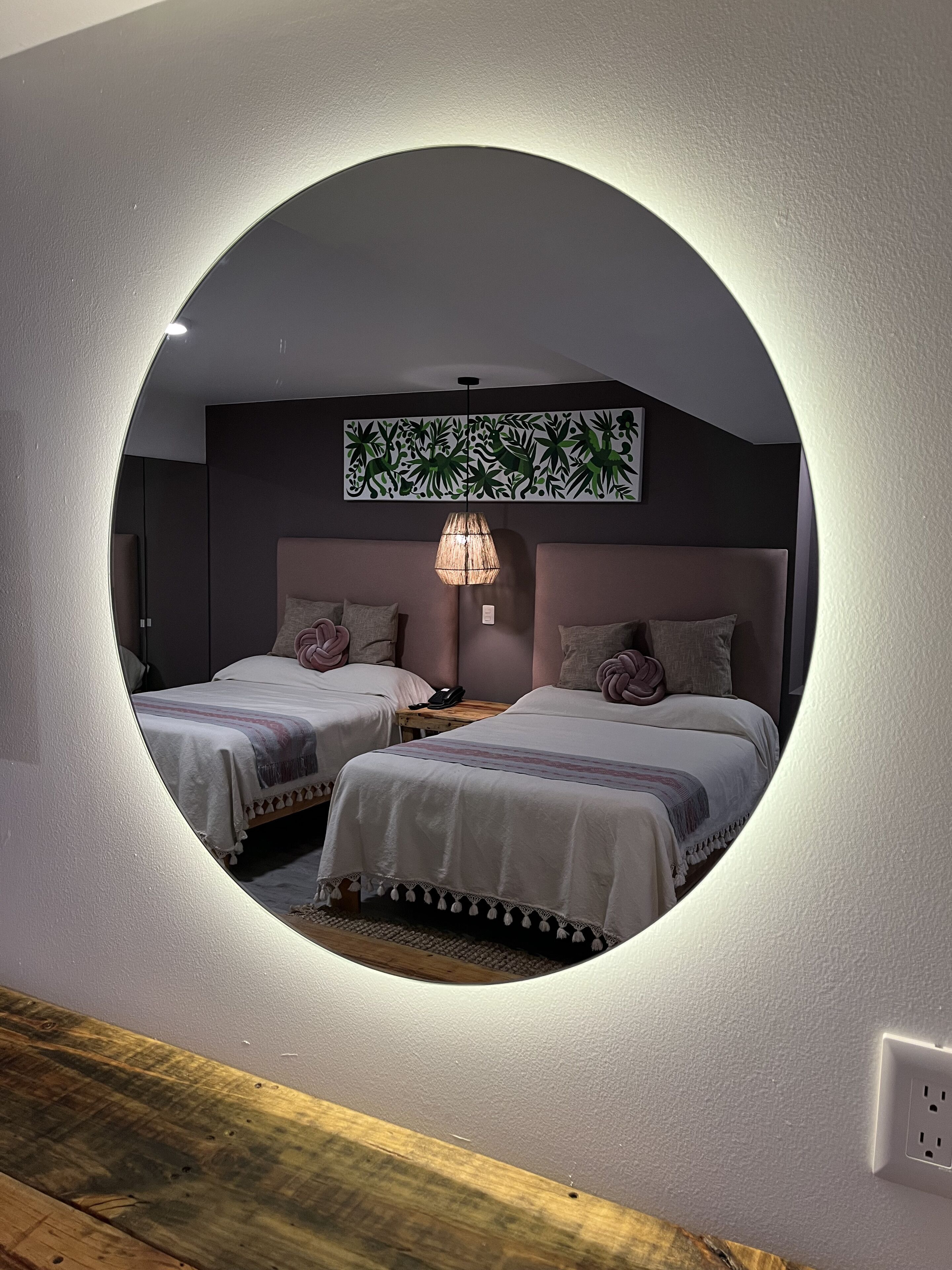 Un espejo circular que refleja un dormitorio con dos camas, una lámpara colgante y una obra de arte en la pared.