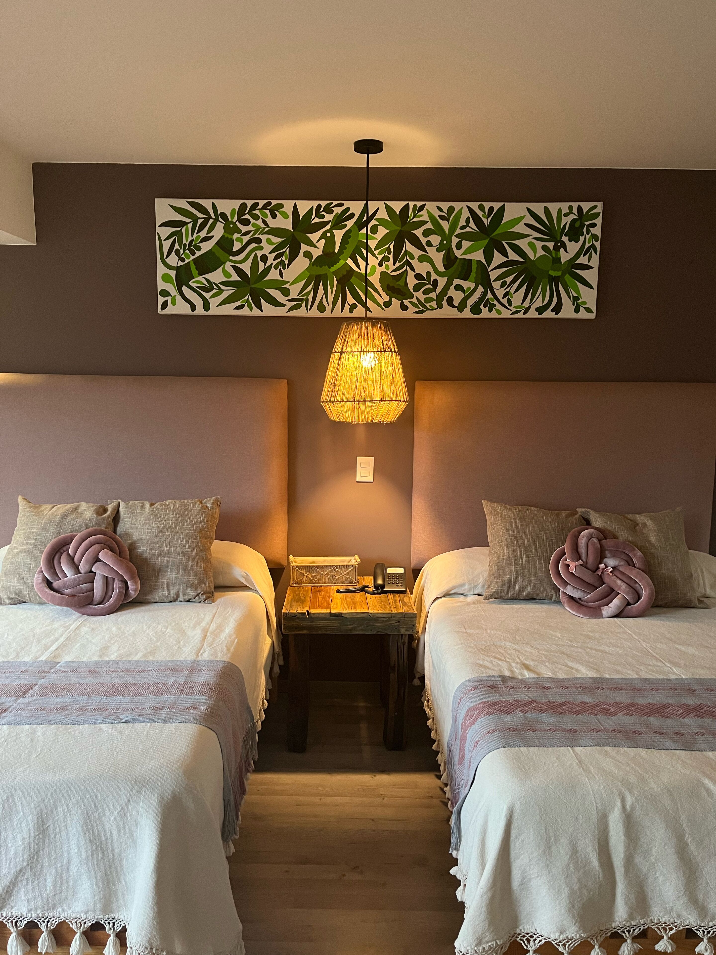 Quarto de hotel com duas camas, luminária pendente trançada, arte floral na parede e uma mesinha com telefone e abajur.