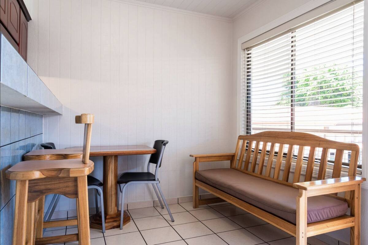 A small, clean room with a wooden table, two chairs, a bench, and a window with blinds.