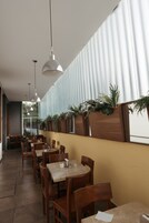 Un restaurante con mesas y sillas de madera, lámparas colgantes y una hilera de macetas a lo largo de la pared.