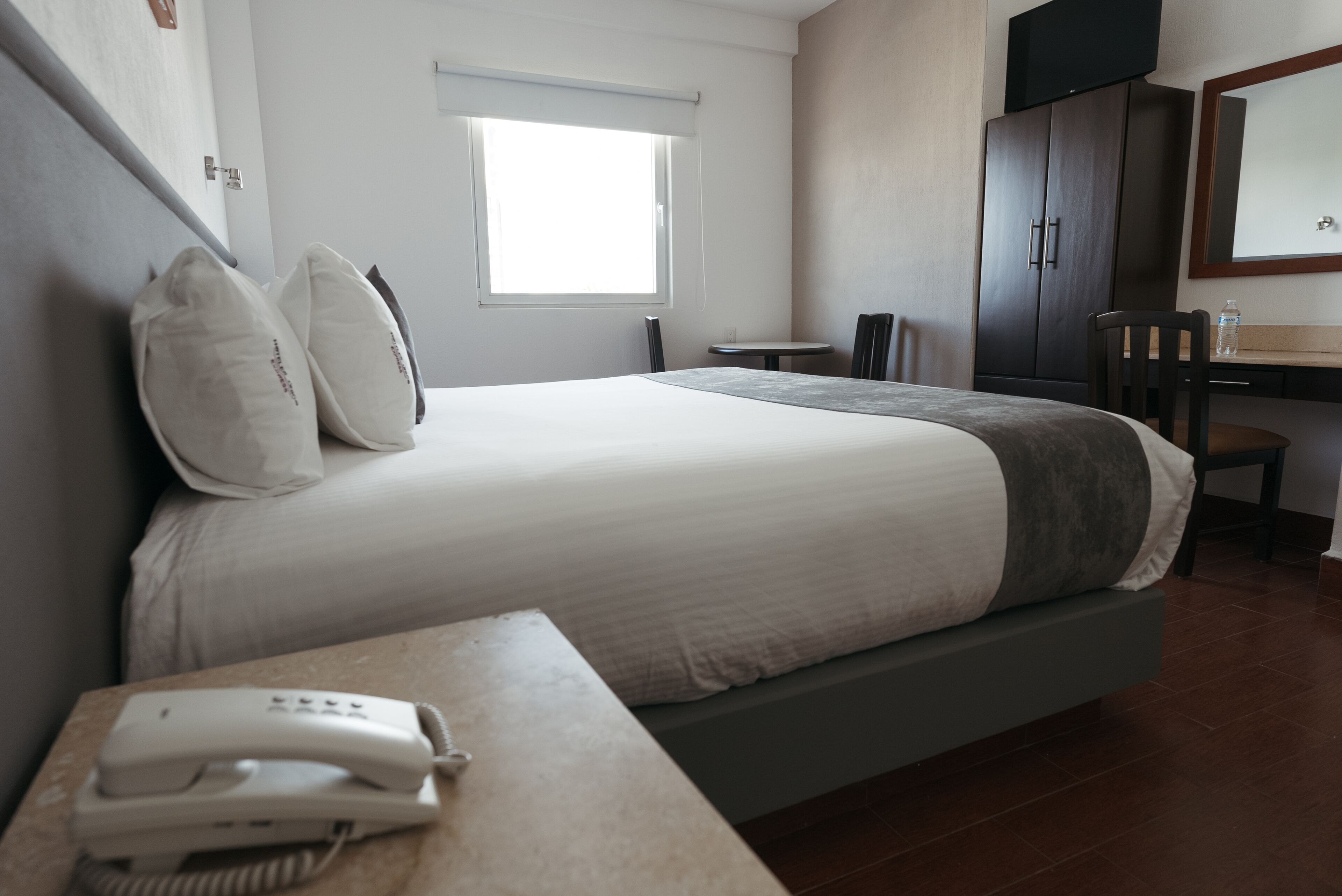Habitación de hotel con cama, mesita de noche con teléfono, una pequeña mesa con sillas, ventana, escritorio con espejo y armario.