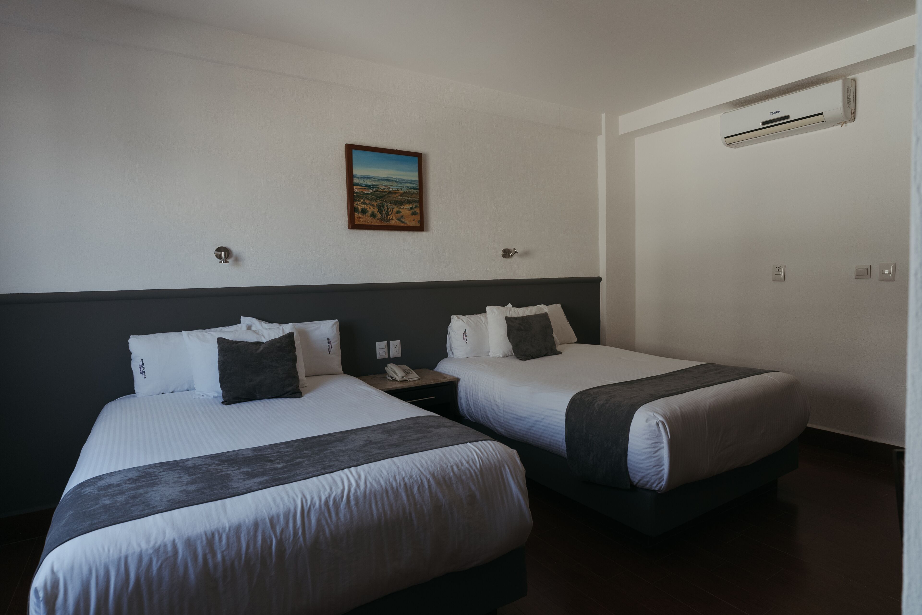 A hotel room with two beds, a painting on the wall, and an air conditioning unit.