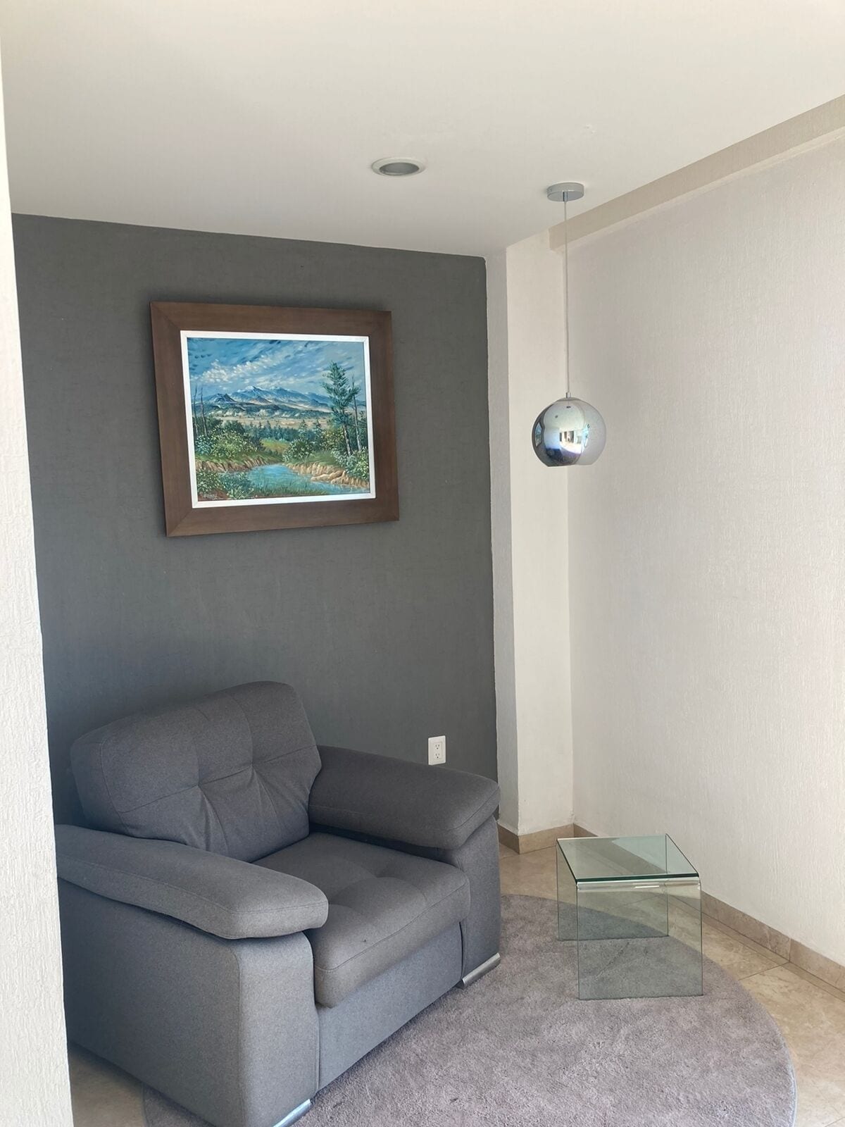 A modern living room with a grey sofa, a framed landscape painting, a glass coffee table, and a wall-mounted light fixture.