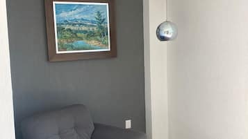 A modern living room with a grey sofa, a framed landscape painting, a glass coffee table, and a wall-mounted light fixture.