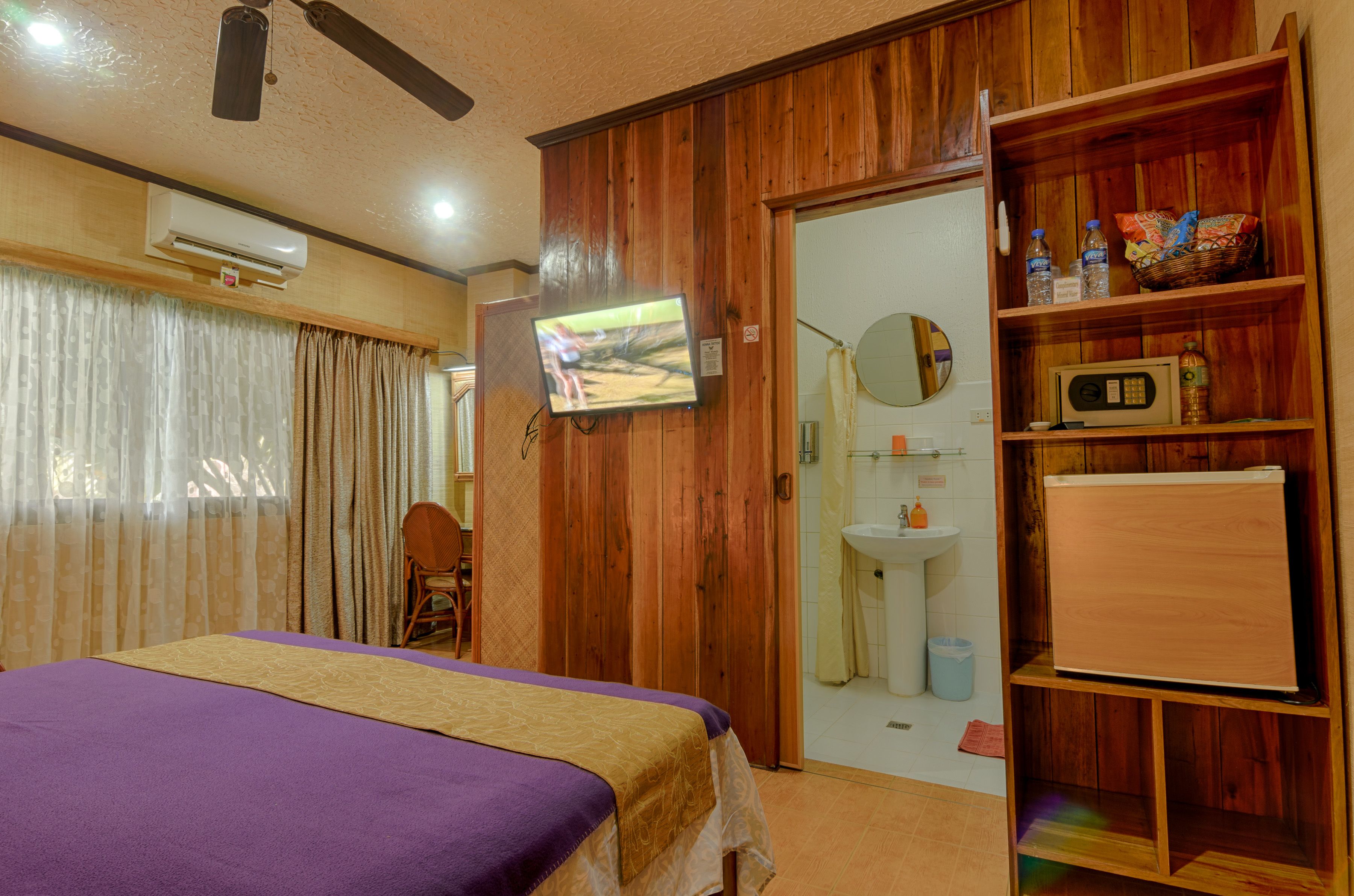 A hotel room with a bed, a television, a wooden wardrobe, a bathroom with a sink and mirror, and an air conditioning unit.