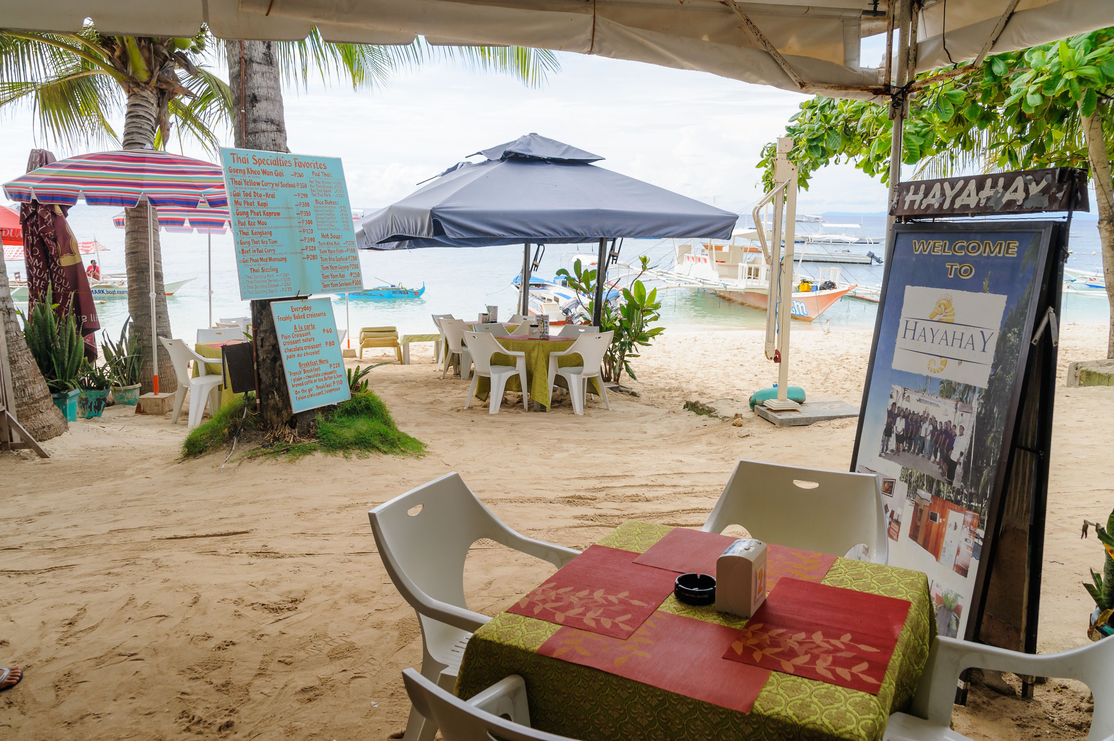 A beachside restaurant with outdoor seating, a menu board, and a welcome sign.