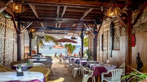 An outdoor dining area with tables and chairs, a view of the sea, and a wooden ceiling with hanging lights.