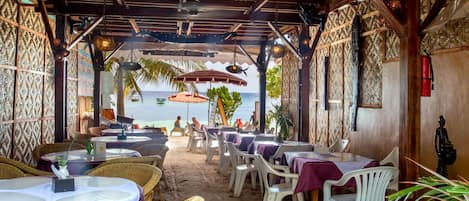 An outdoor dining area with tables and chairs, a view of the sea, and a wooden ceiling with hanging lights.