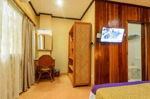 A hotel room with a bed, a television mounted on the wall, a wooden wardrobe, a desk with a chair, and a bathroom visible through an open door.
