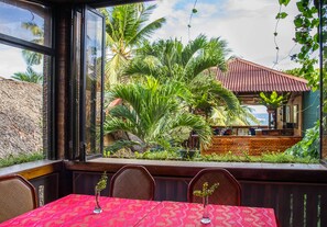 An outdoor dining area with a red patterned tablecloth, wicker chairs, and a view of a building with a red roof and palm trees. - Hayahay Resort (Panglao)