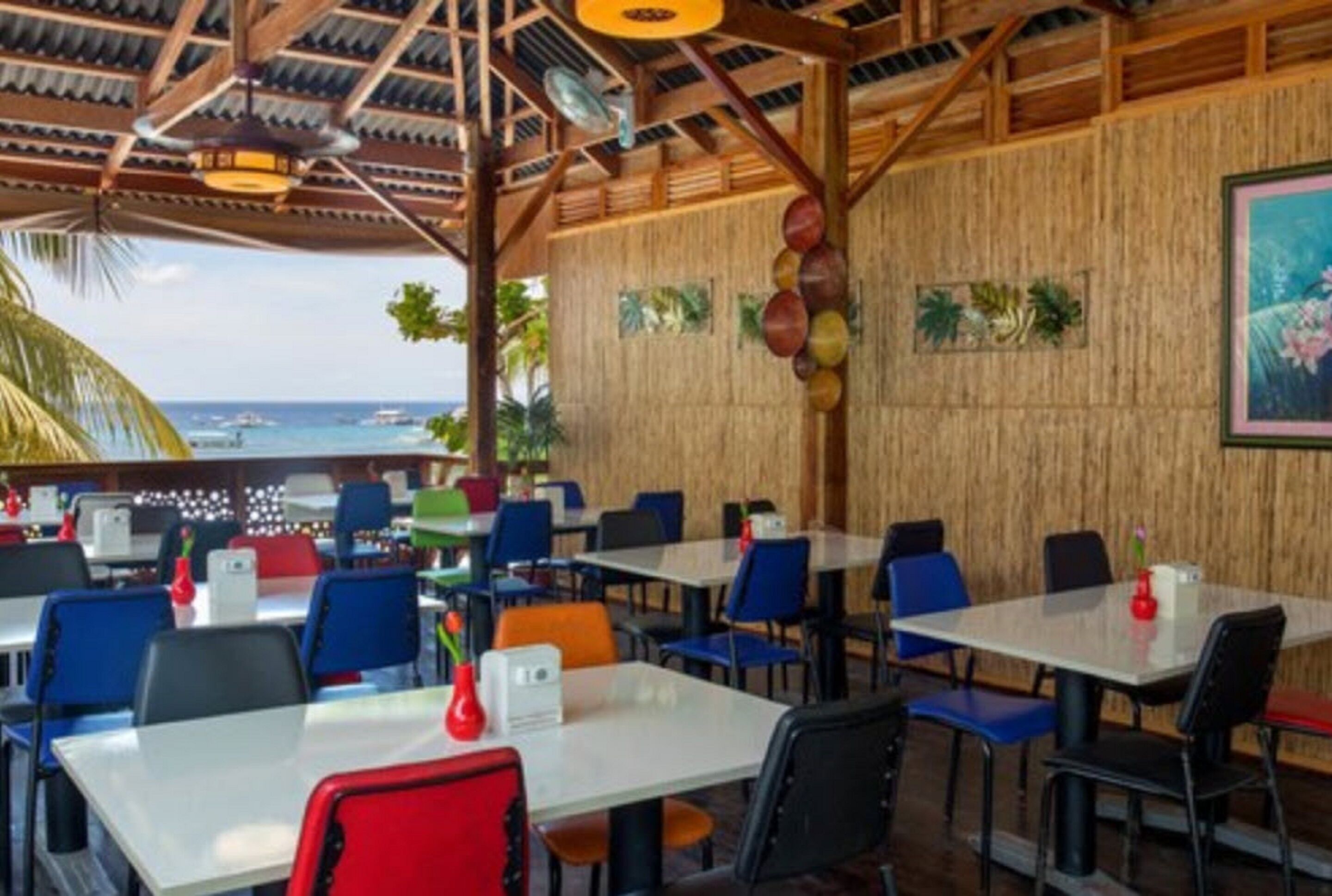 A restaurant with a view of the ocean, featuring tables and chairs, wooden walls, and tropical decor.