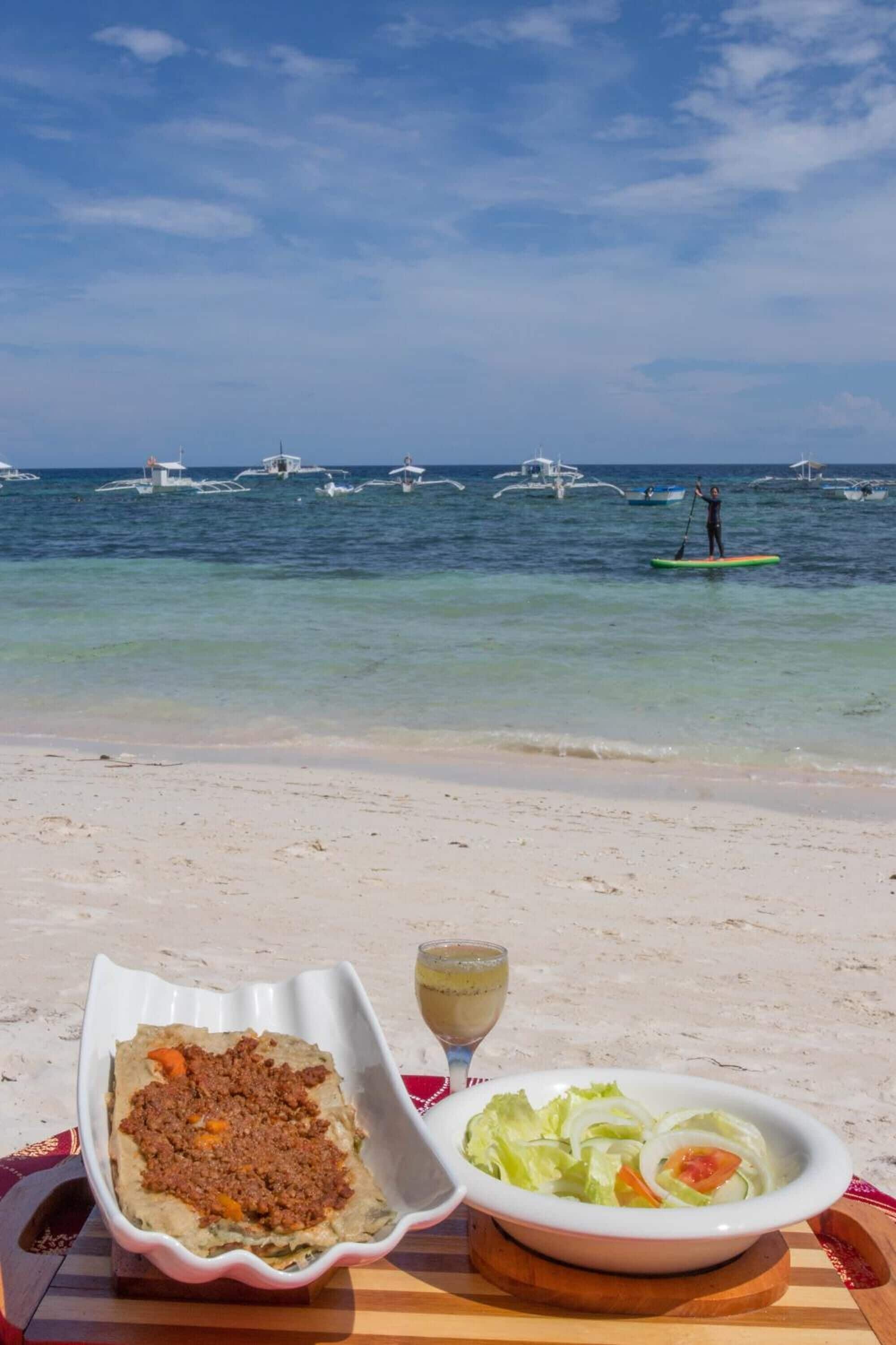 A beach scene with a table set with food and a drink, boats in the background.