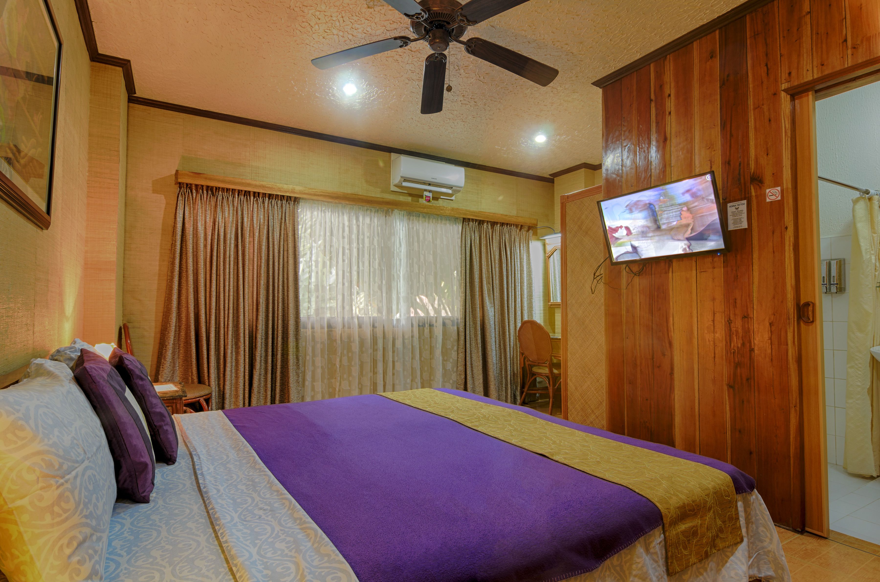Une chambre d’hôtel avec un lit, un ventilateur de plafond, une télévision et une porte en bois.