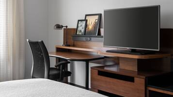 A hotel room with a wooden desk, a black office chair, a television, and a bed.