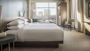 A modern hotel room with a large bed, a seating area with a sofa and armchairs, a side table with a vase, and a large window offering a city view.