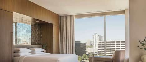 A modern hotel room with a large bed, a seating area, and a view of the cityscape through large windows.