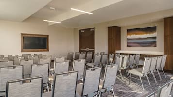 Une salle de conférence avec des rangées de chaises grises, un grand écran et un tableau accroché au mur.
