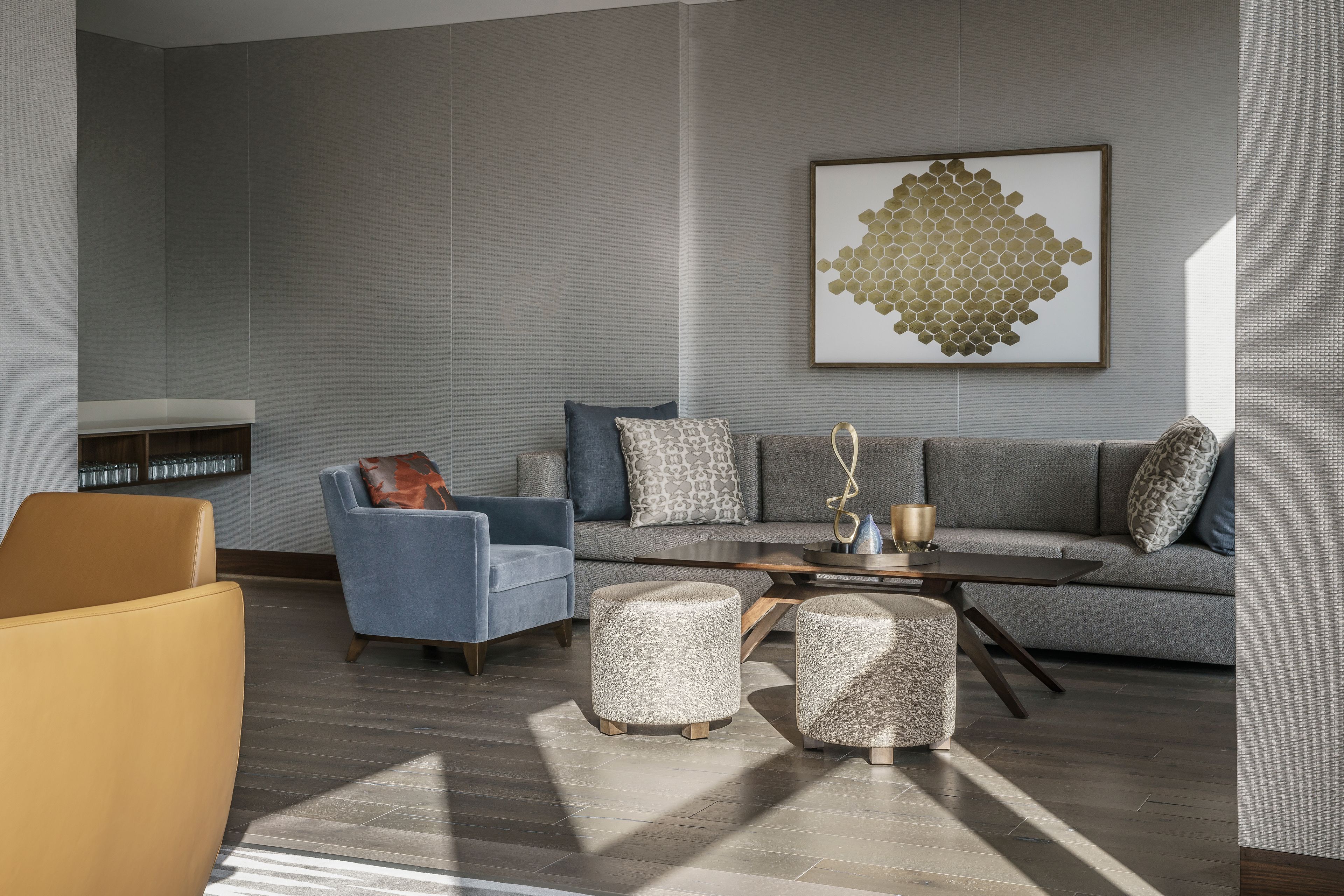 A modern living room with a grey sofa, blue armchair, and ottomans. A coffee table and a framed artwork are on the sofa.