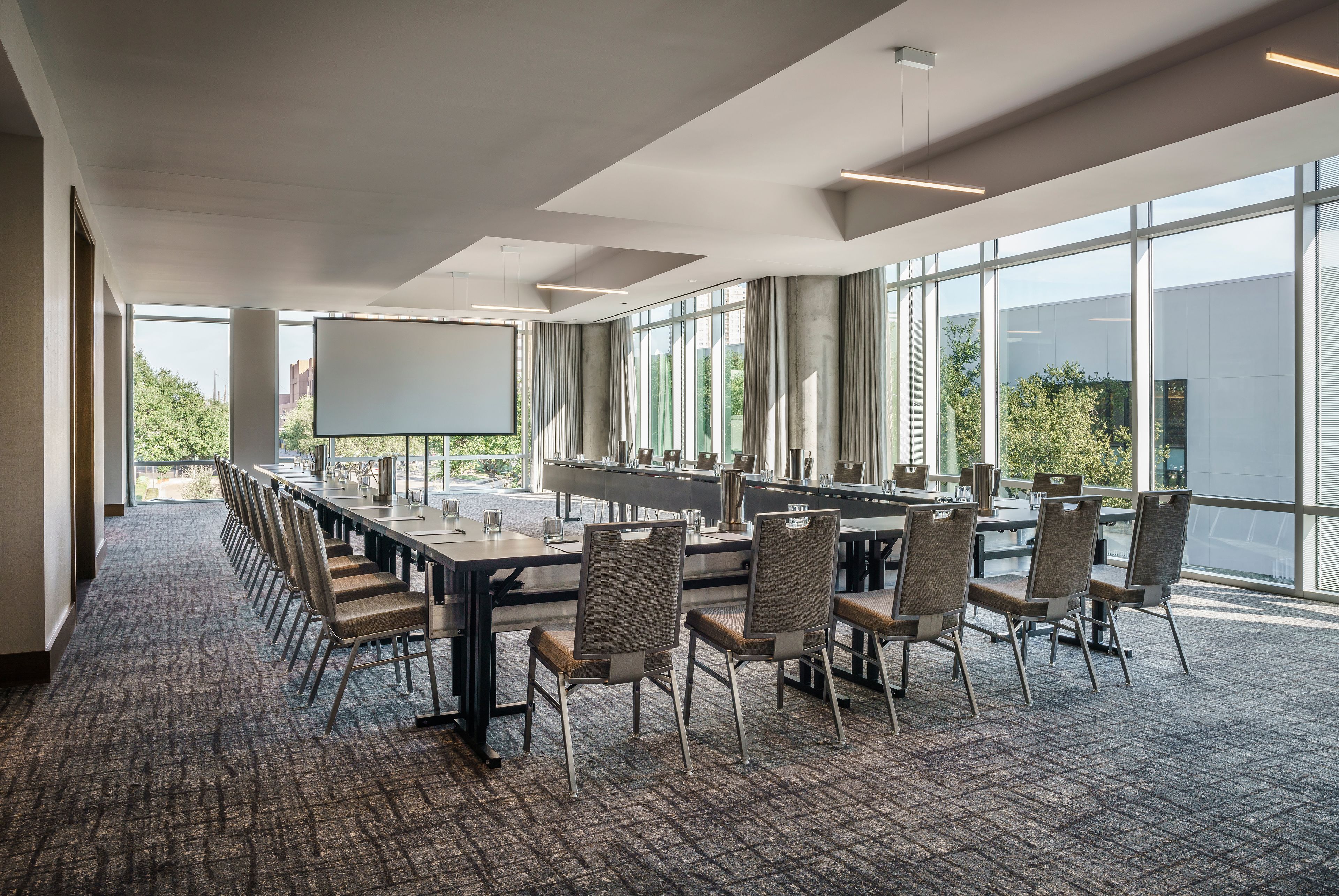 Un salón de conferencias con una larga mesa, sillas, una pantalla de proyector y ventanas amplias que dan a vistas de follaje exterior.