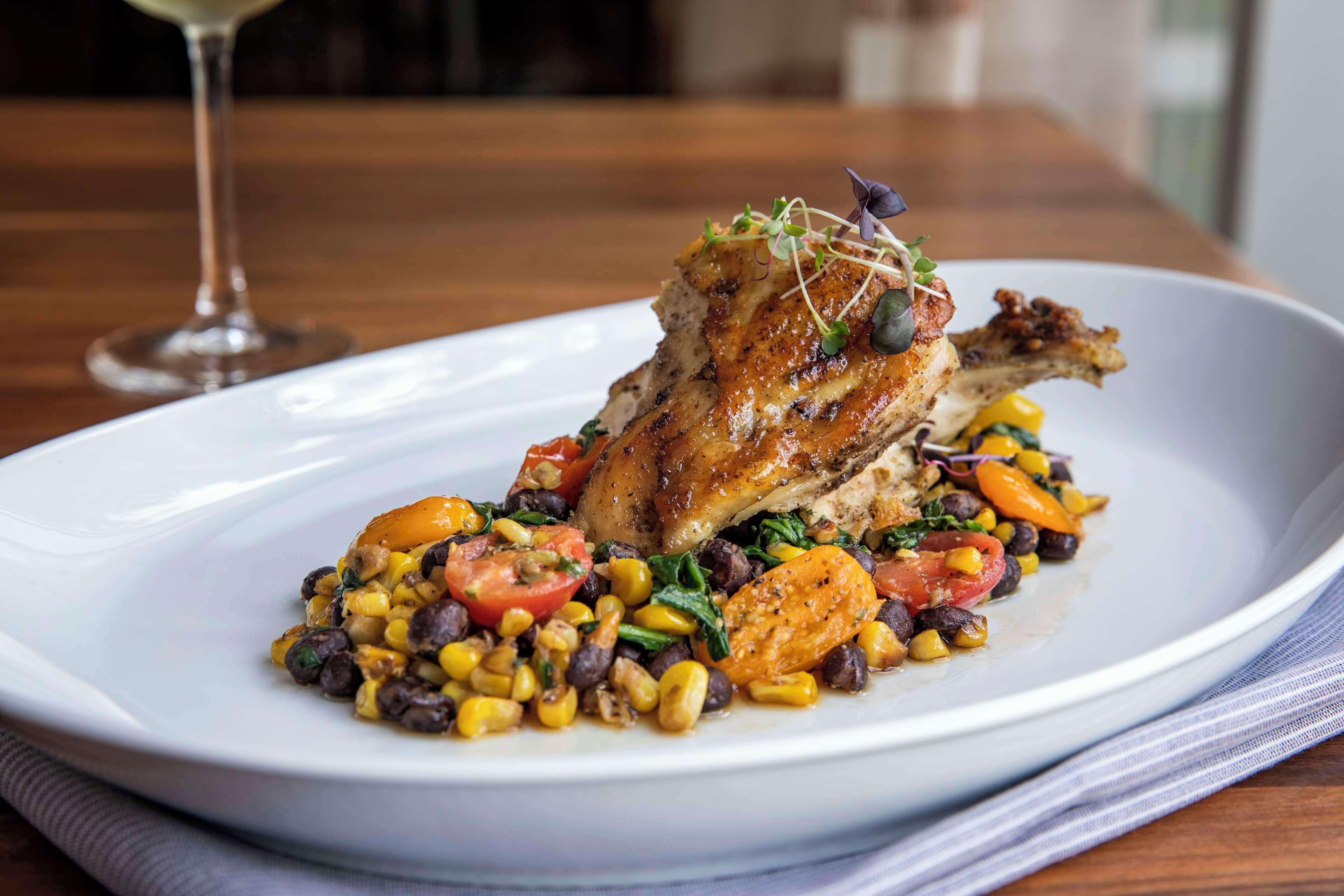 A plate of food with a roasted chicken leg, vegetables, and a glass of wine on a wooden table.