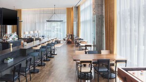 A modern bar area with a long counter, bar stools, and a television screen. There are tables and chairs arranged for dining, and large windows with curtains.