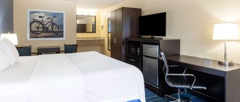 A hotel room with a large bed, a desk with a chair, a television, a refrigerator, and a bathroom with a mirror and sink.