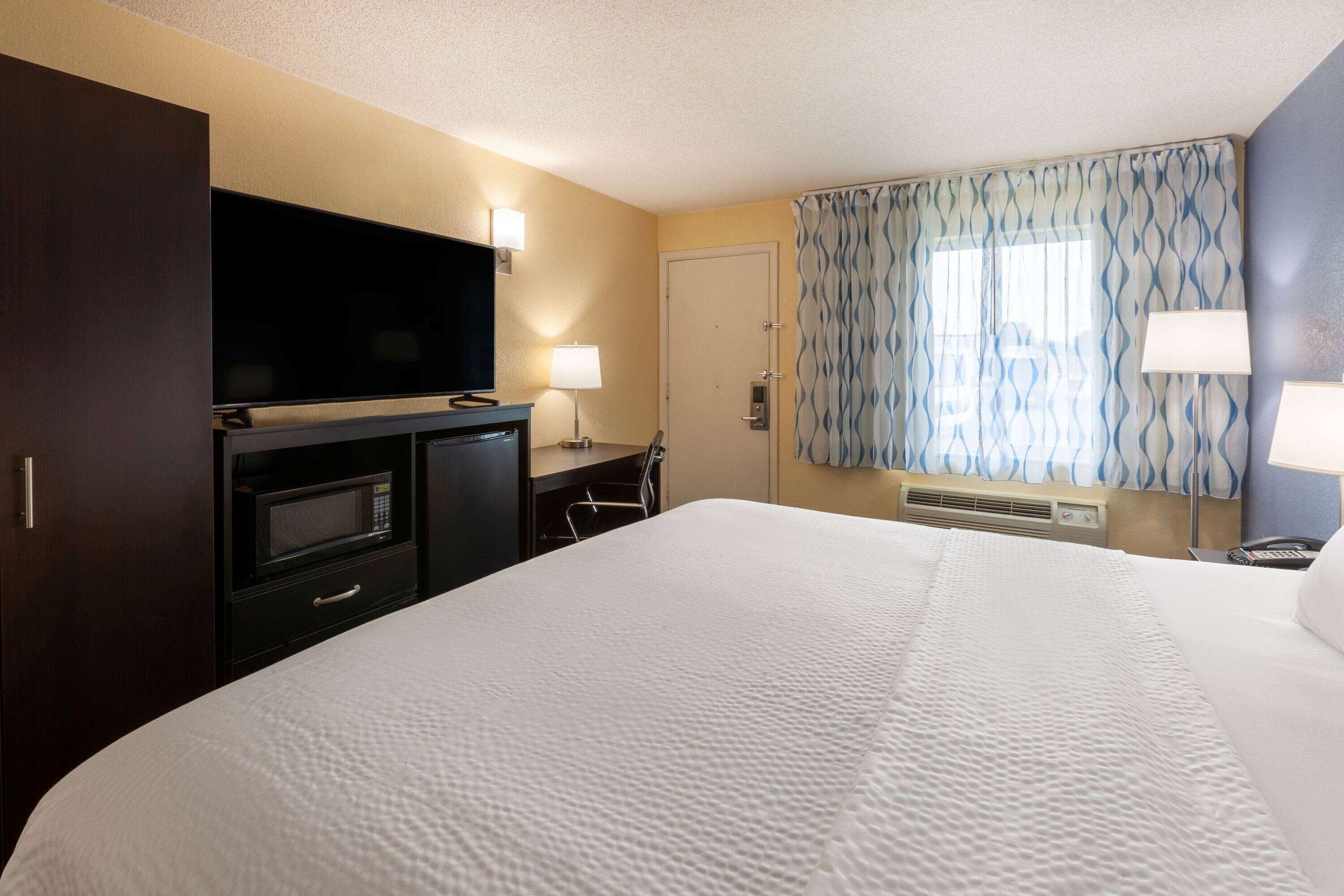 A hotel room with a large bed, a flat-screen TV, a microwave, a desk with a lamp, and a window with curtains.