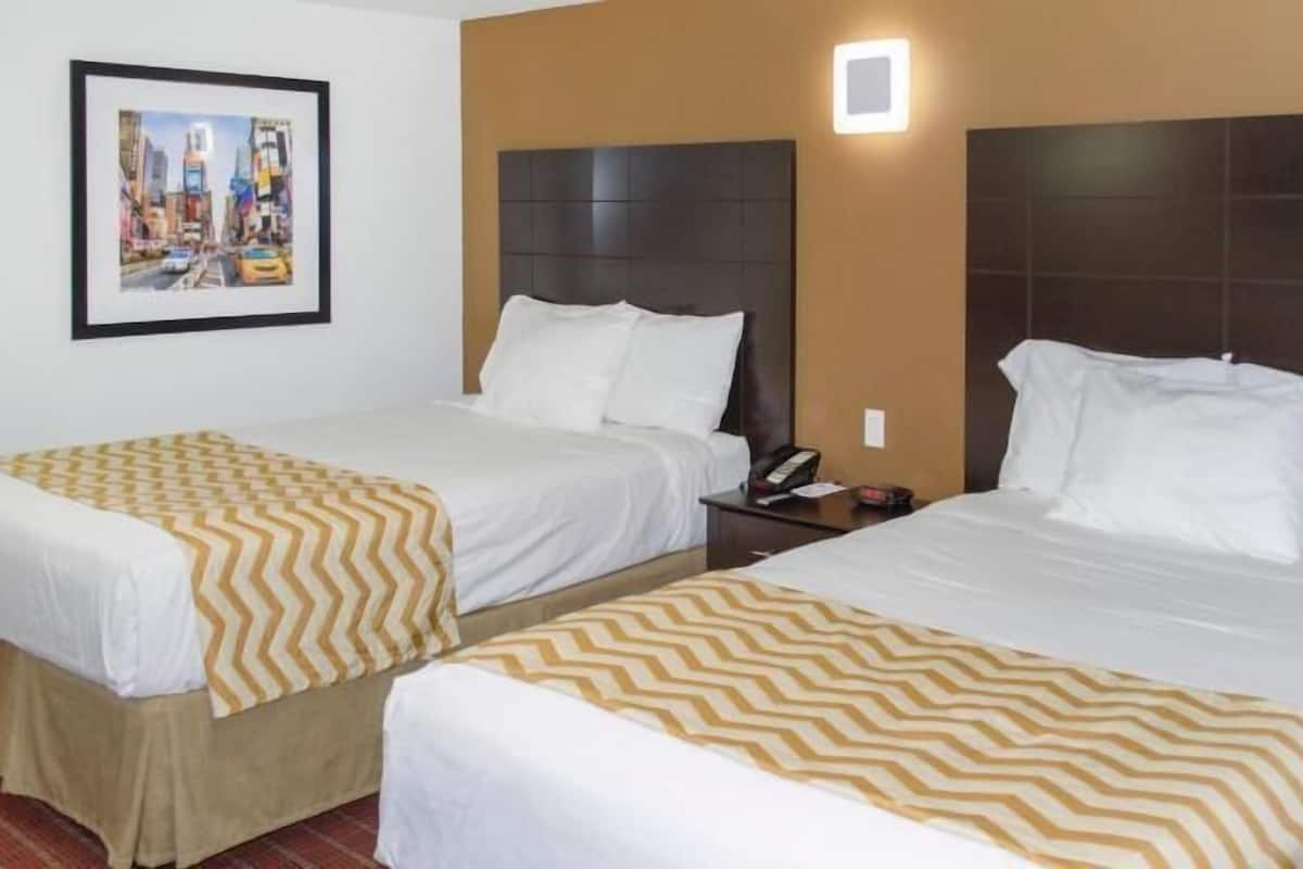 Une chambre d’hôtel avec deux lits, chacun agrémenté de draps blancs et d’un couvre-lit à motifs. Un tableau encadré orne le mur, et une table de chevet avec un téléphone se trouve à côté de chaque lit.