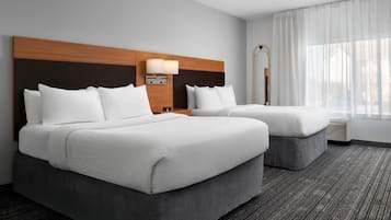 A hotel room with two beds, a wooden headboard, a nightstand with a lamp, a mirror, and a window with sheer curtains.