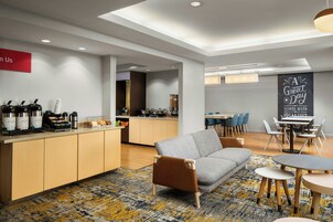 Quarto de hotel moderno com cozinha compacta, um sofá cinza, uma área de jantar e uma placa com a inscrição 'Um Bom Dia Começa Com o Café da Manhã'.