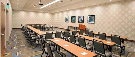 Un salón de conferencias con varias mesas y sillas, un proyector y cuadros en la pared.